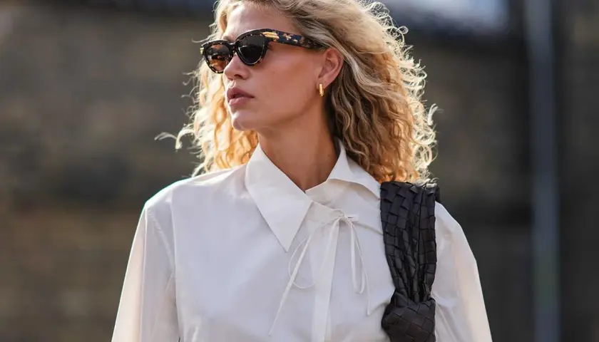 Eine skandinavische Blonde mit lockigem Haar, Sonnenbrille und weißem Hemd, die eine dunkle Tasche trägt.