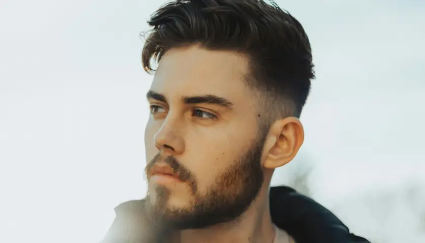 Junger Mann mit Bart und einem modernen taper fade cut, der nachdenklich in die Ferne blickt.