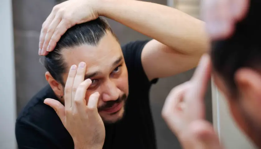 Mann prüft seine Haare im Spiegel. Er überlegt, ob Aminexil oder Minoxidil besser gegen Haarausfall hilft.