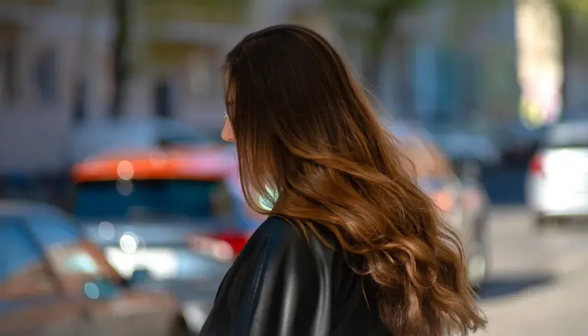 Braune Haare mit Beach Waves, die im Wind wehen. Die Frau trägt eine schwarze Lederjacke. Im Hintergrund verschwommene Autos.