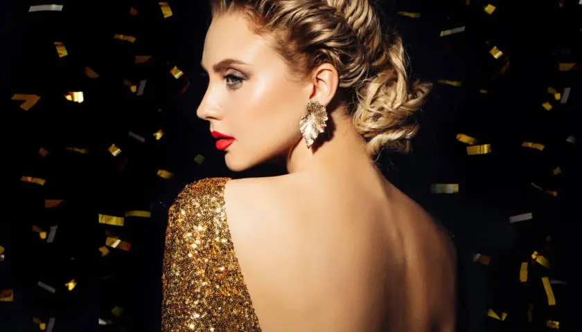 Elegante Frau mit aufwendiger Hochsteckfrisur, perfekt für Silvester-Frisuren. Goldglitzerndes Kleid und Konfetti im Hintergrund.