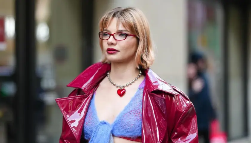 Frau mit roter Brille und einem roten Herz-Anhänger trägt eine glänzende rote Jacke über einem blauen Strickoberteil. Ihr **bob hinten kurz gestuft** umrahmt ihr Gesicht.