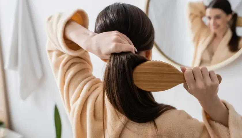 Frau bürstet ihr langes, dunkles Haar mit einer Holzbürste. Sie trägt einen Bademantel. Ein Spiegel zeigt sie lächelnd. So kann man **Haare schneller wachsen lassen**.