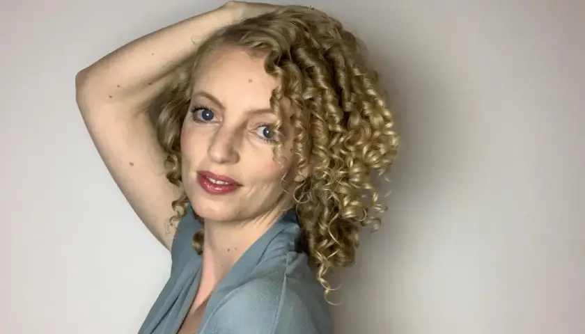 Frau mit blonden, voluminösen 3a locken, die ihren Arm über den Kopf hebt.
