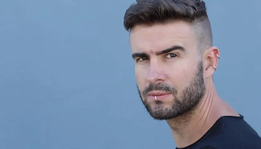 Junger Mann mit kurz geschorenen Seiten und Haare nach hinten, Bart und Lippenpiercing.