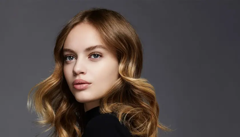 Porträt einer jungen Frau mit welligem, dunkelblondem Balayage-Haar, das sanft um ihr Gesicht fällt.
