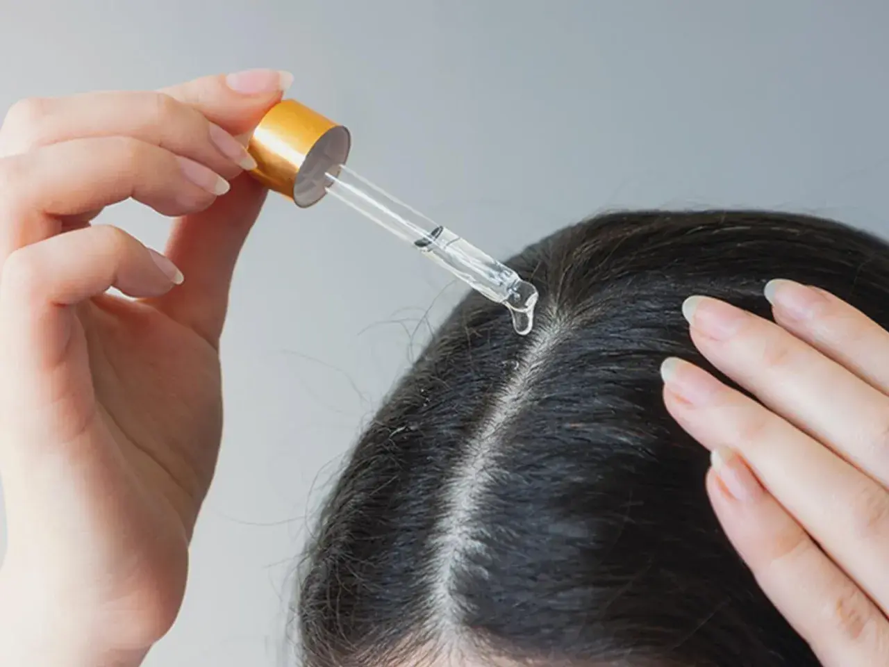 Eine Hand trägt mit einer Pipette eine Flüssigkeit auf die Kopfhaut auf. Ein Tropfen fällt auf den Scheitel. Apfelessig gegen Schuppen kann hier helfen.
