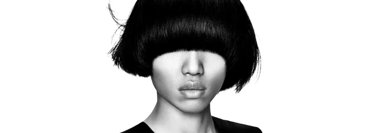Ein Gesicht mit einem markanten **bowl cut** und dunklem Haar, das die Augen verdeckt. Die Lippen sind leicht geöffnet.
