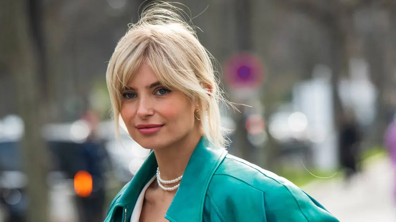 Blonde Frau mit soft curtain bangs, trägt eine türkise Lederjacke und Perlenkette.