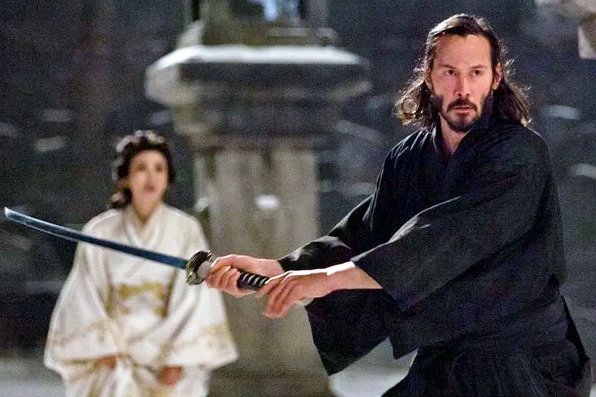Ein Mann mit langer, dunkler Samurai-Frisur und Bart hält ein Schwert. Im Hintergrund eine Frau in weißem Kimono.