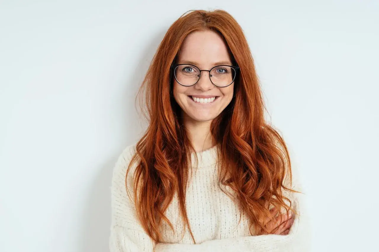 Eine lächelnde Frau mit langen, roten Haaren und Brille. Ihr Outfit, ein cremefarbener Strickpullover, steht ihr gut.