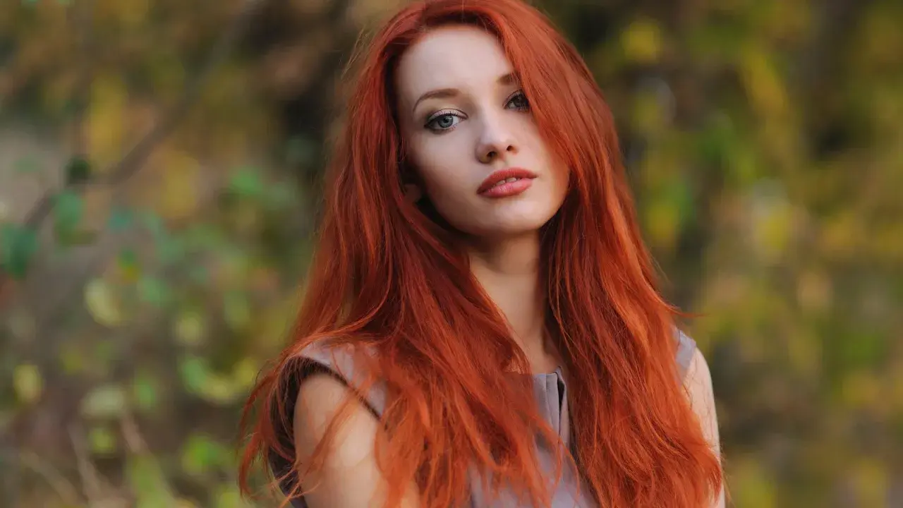 Eine Frau mit langen, dunkelroten Haaren blickt nachdenklich in die Kamera. Ihr Haar fällt ihr über die Schultern, während sie im Freien steht.
