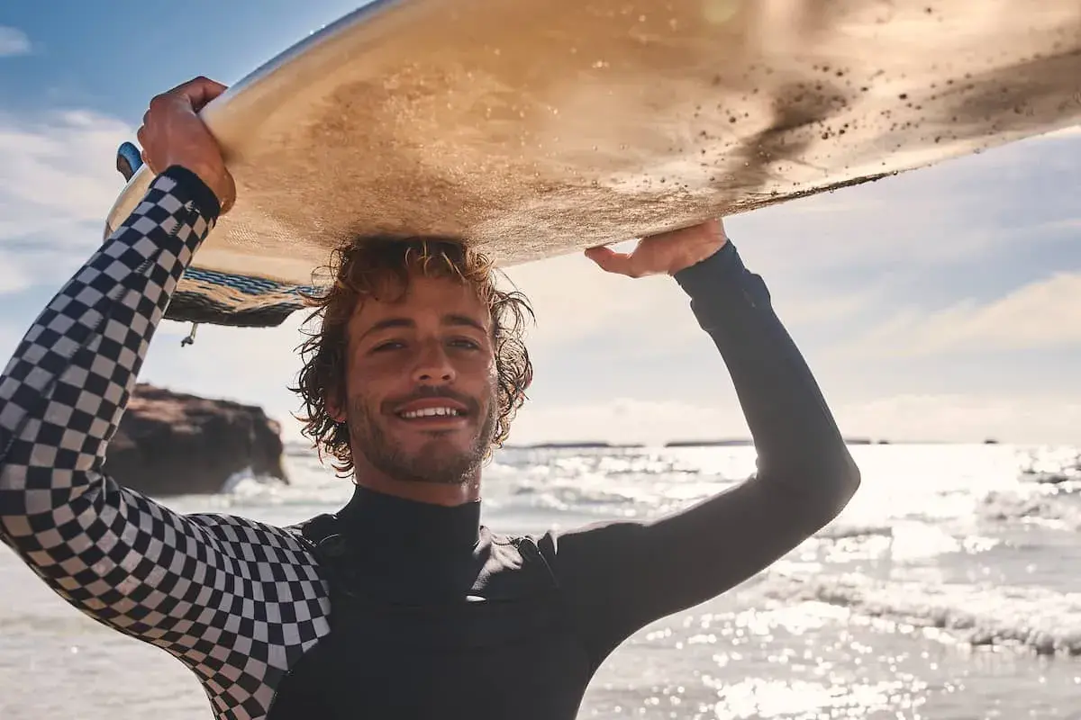 Lächelnder Surfer mit Wellen im Haar trägt sein Brett über dem Kopf. Sein Neoprenanzug hat ein cooles Schachbrettmuster.