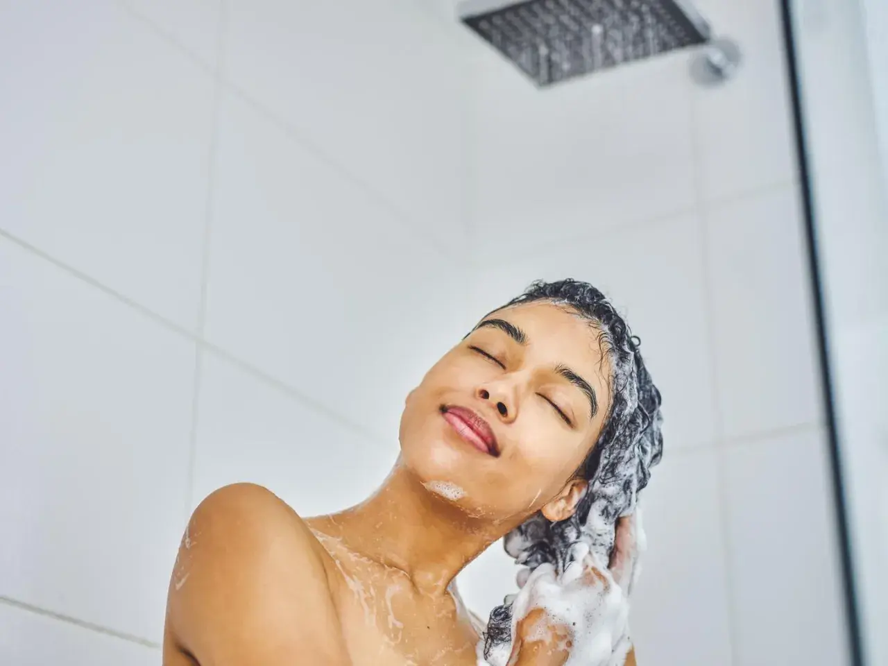 Frau genießt eine tiefenreinigende Haarwäsche unter der Dusche. Schaumige Hände massieren das Shampoo ins nasse Haar.