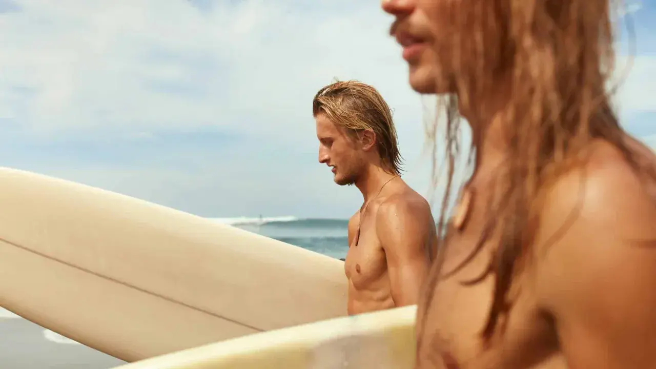 Zwei Surfer am Strand, einer mit einer Surfer Frisur kurz, tragen Surfbretter.