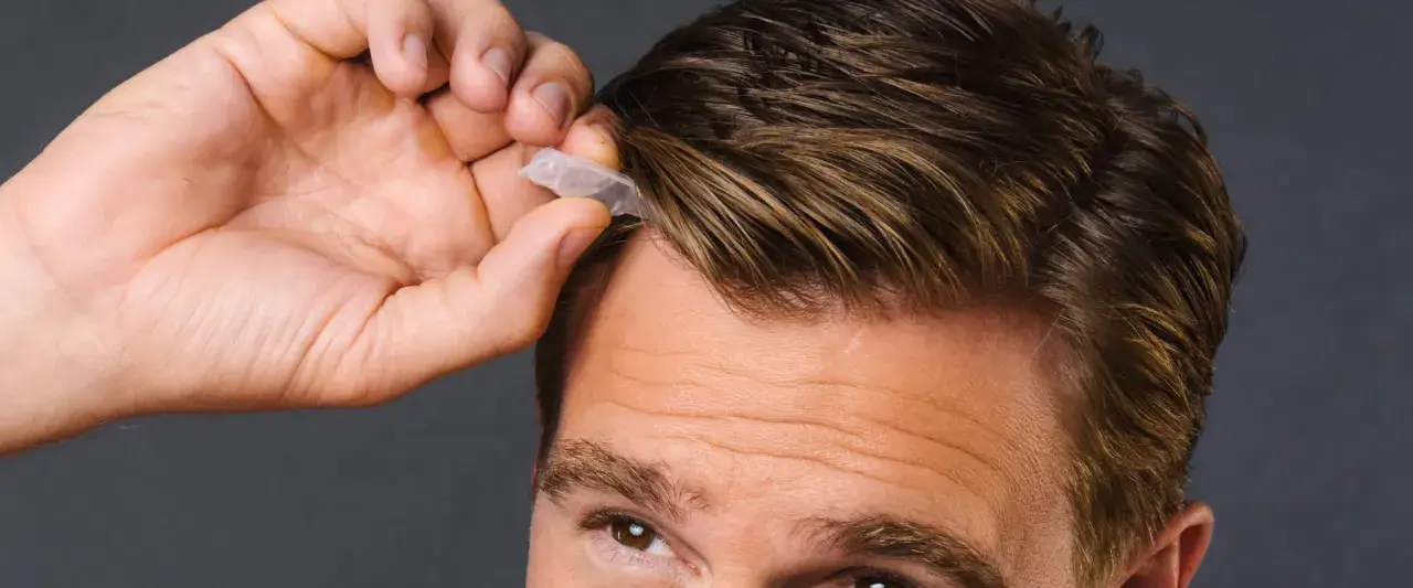 Mann mit beginnenden Geheimratsecken, der eine kleine Kapsel ins Haar drückt.