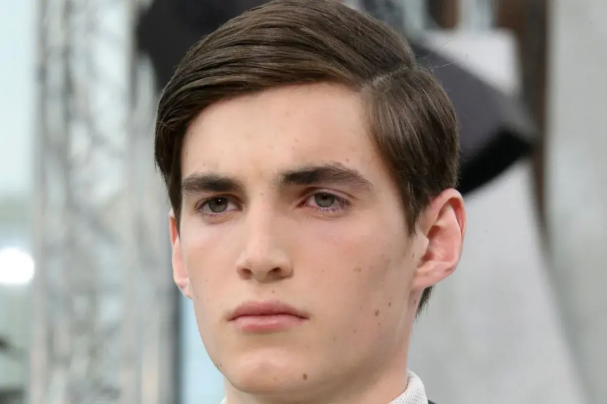 Ein junger Mann mit braunem Haar, das zu einem perfekten Seitenscheitel gestylt ist. Sein Blick ist nachdenklich.