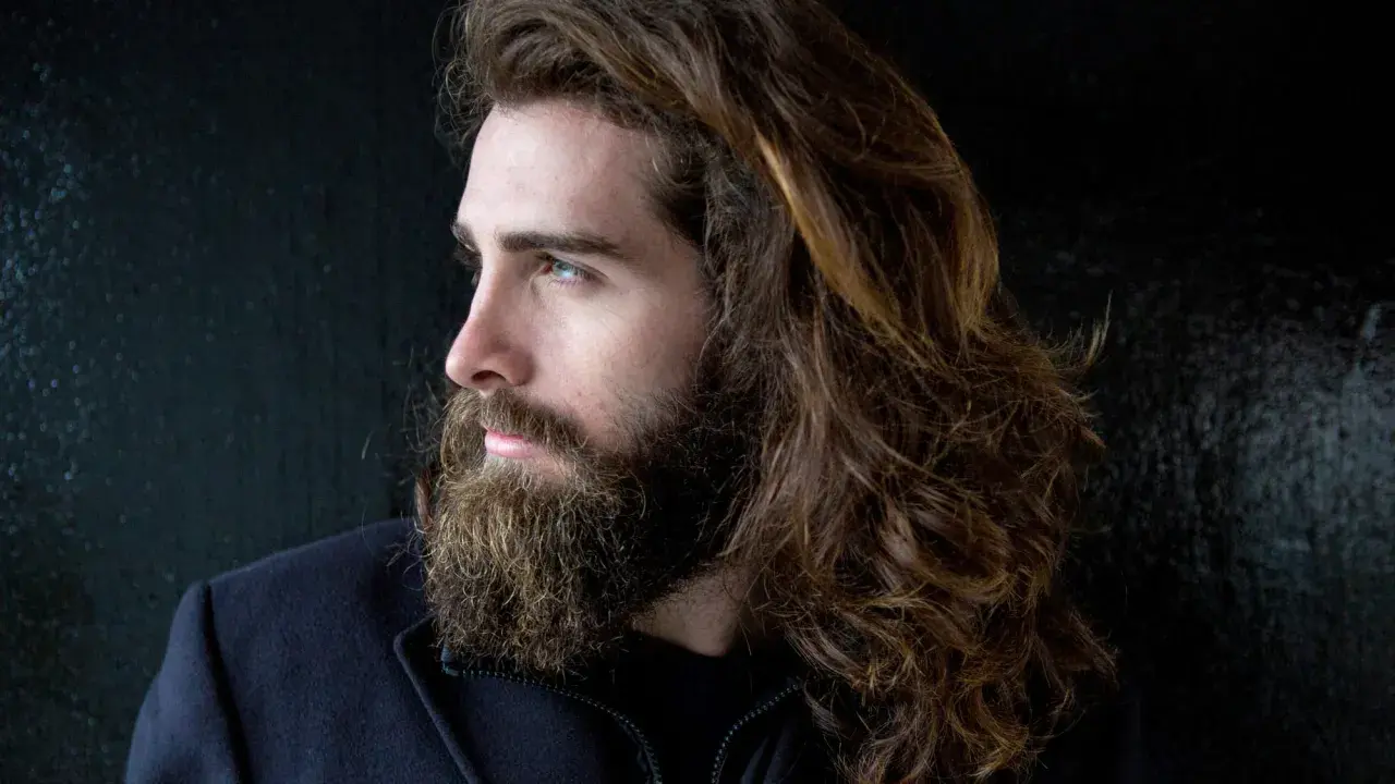 Mann mit langen, braunen Haaren und Bart, der stolz seine Entscheidung zeigt, die Haare wachsen zu lassen.