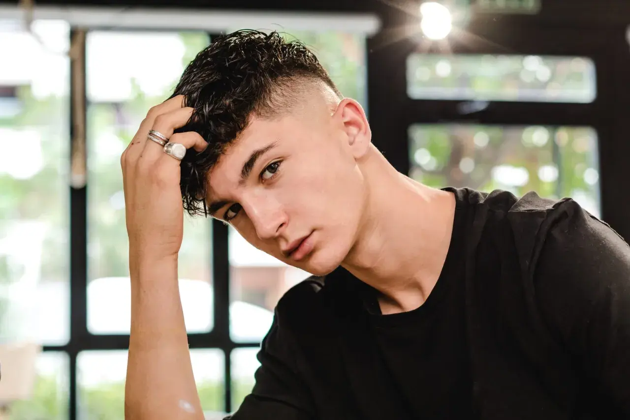 Junger Mann mit kurzgeschorenen Seiten und einem kurzen Mullet, der nachdenklich seine Hand an den Kopf legt.
