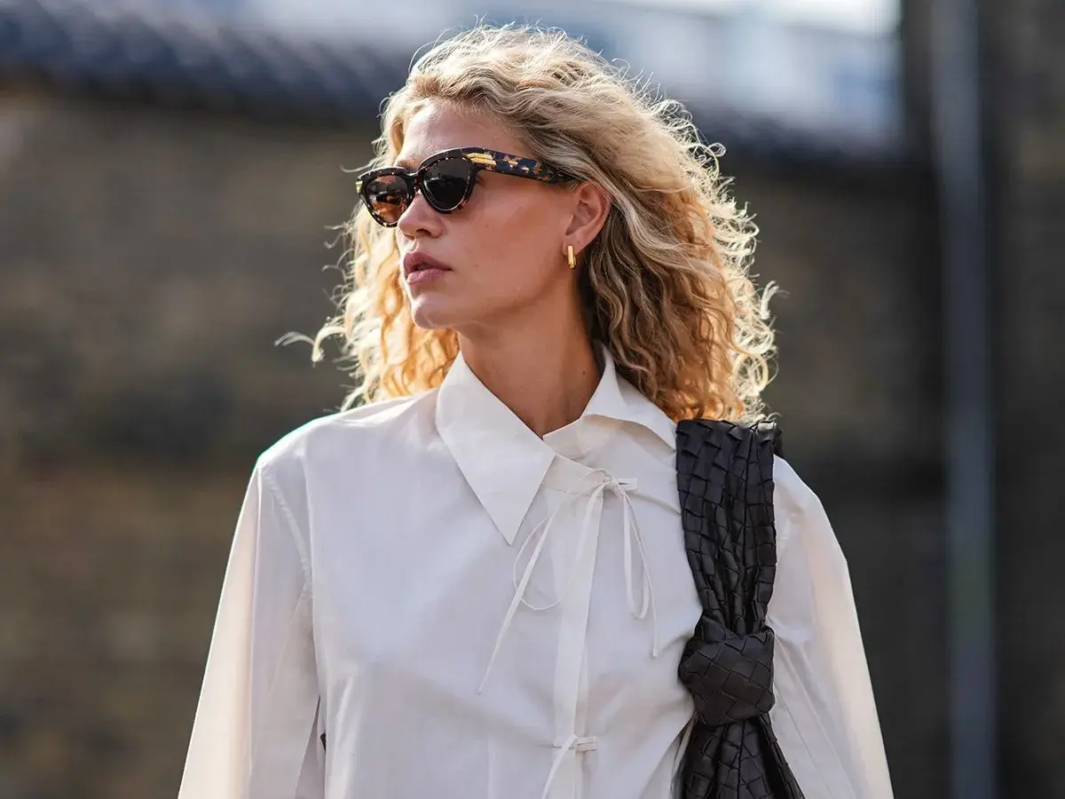 Eine skandinavische Blonde mit lockigem Haar, Sonnenbrille und weißem Hemd, die eine dunkle Tasche trägt.