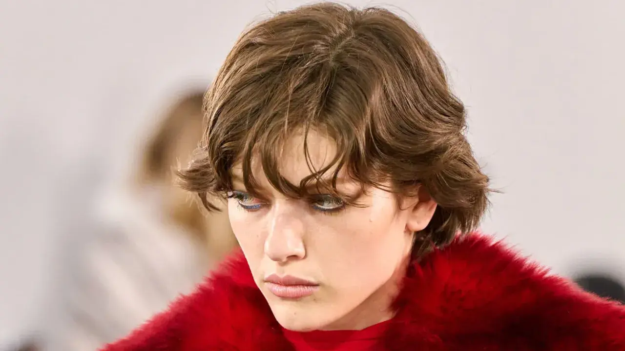 Ein Model mit modernen Kurzhaarfrisuren und auffälligem blauen Eyeliner, gekleidet in einen roten Pelzmantel.