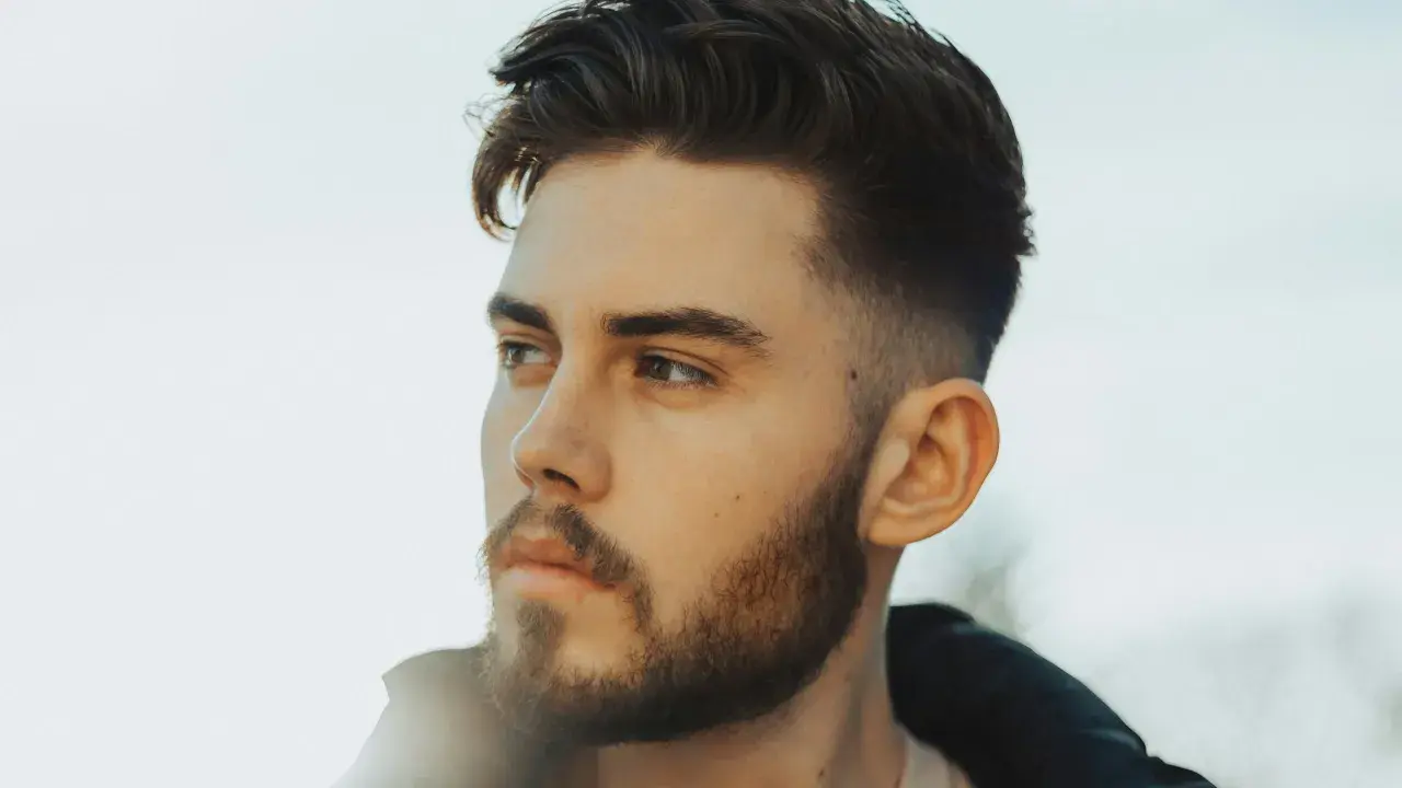 Junger Mann mit Bart und einem modernen taper fade cut, der nachdenklich in die Ferne blickt.