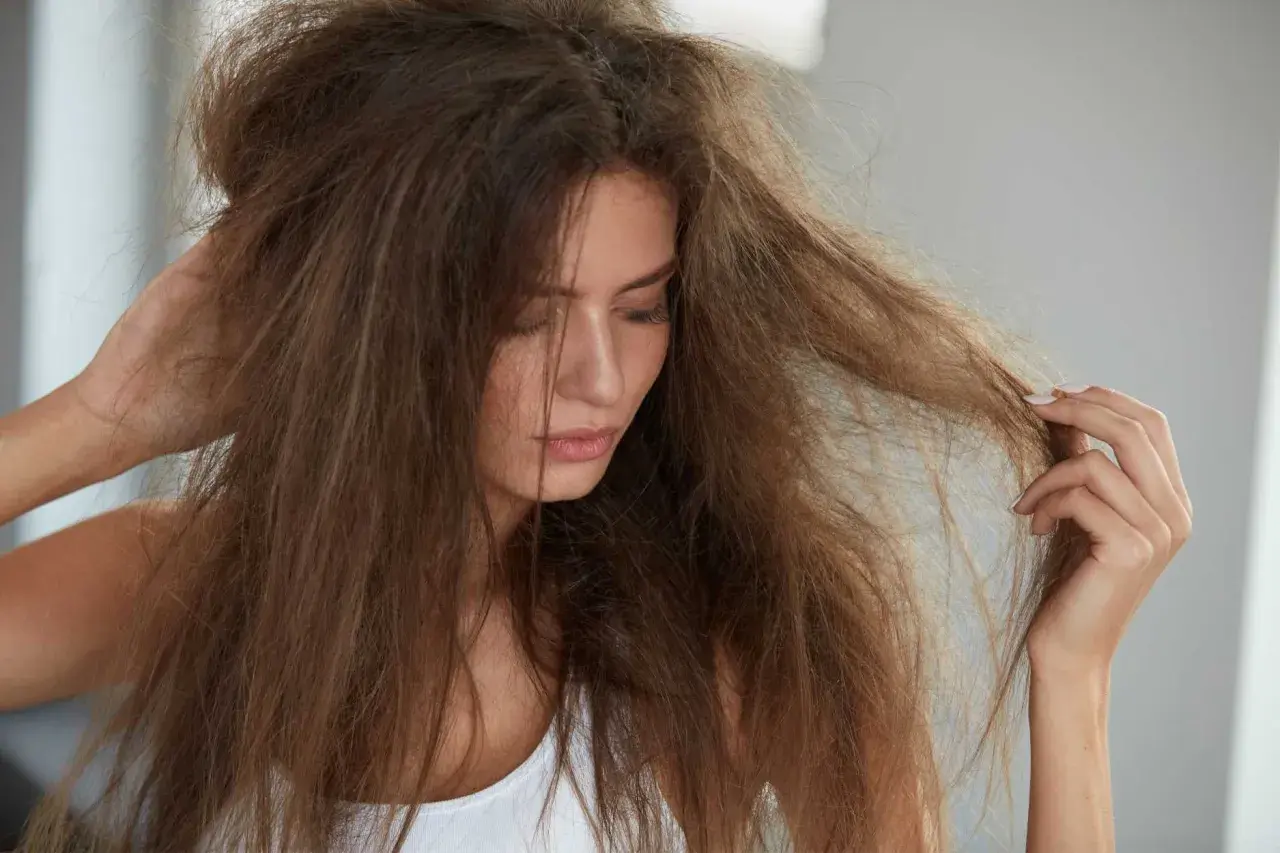 Frau mit zerzaustem Haar, das sich trocken anfühlt. Ihre trockene Haarspitzen sind sichtbar, als sie nachdenklich ihr Haar berührt.