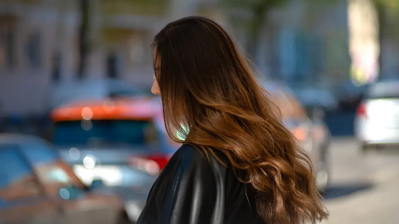 Braune Haare mit Beach Waves, die im Wind wehen. Die Frau trägt eine schwarze Lederjacke. Im Hintergrund verschwommene Autos.