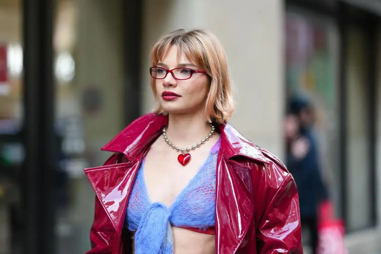 Frau mit roter Brille und einem roten Herz-Anhänger trägt eine glänzende rote Jacke über einem blauen Strickoberteil. Ihr **bob hinten kurz gestuft** umrahmt ihr Gesicht.