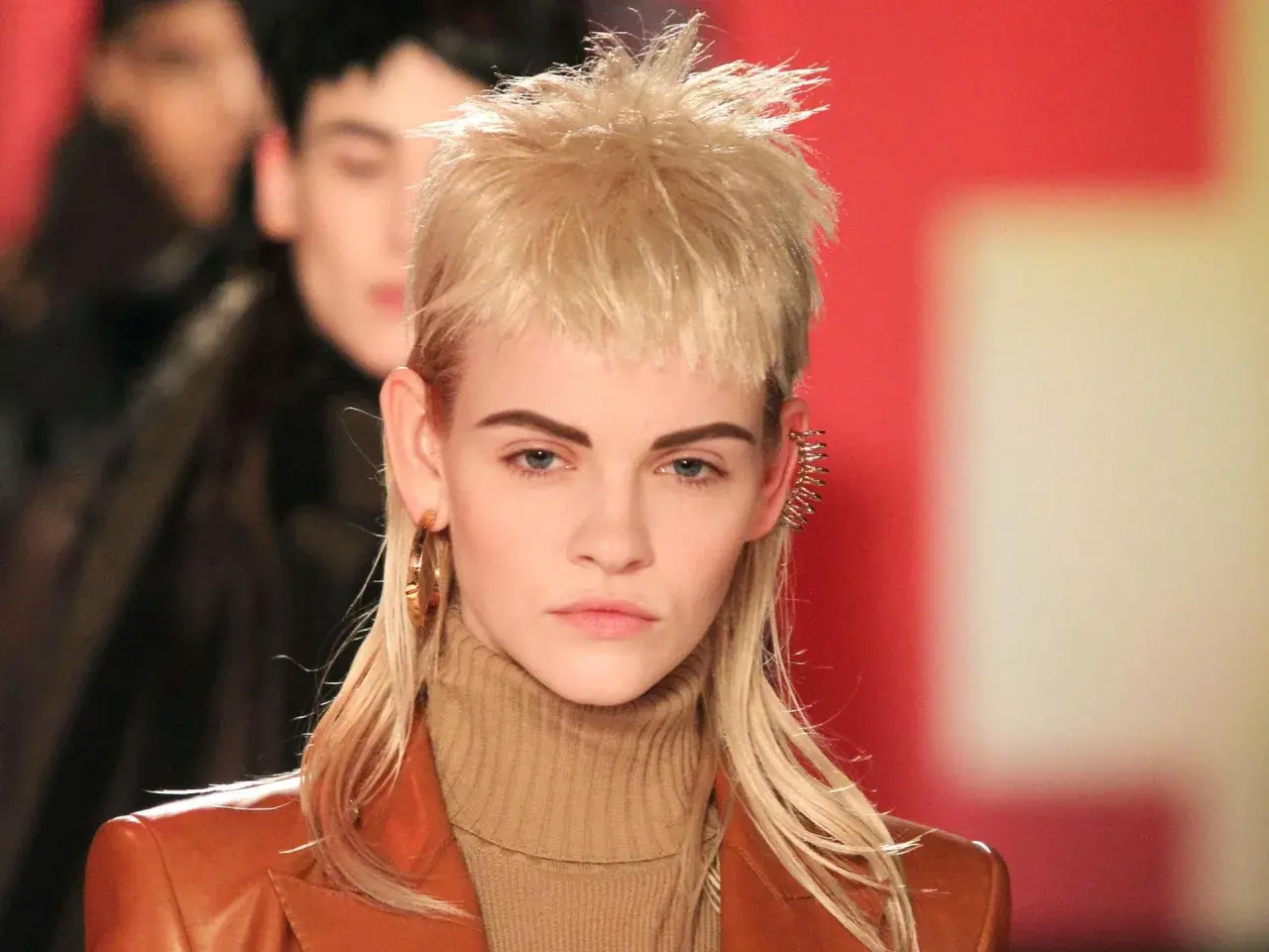 Ein Model präsentiert einen modernen Mullet mit blonden Locken, auffälligen Ohrringen und einem braunen Rollkragenpullover.