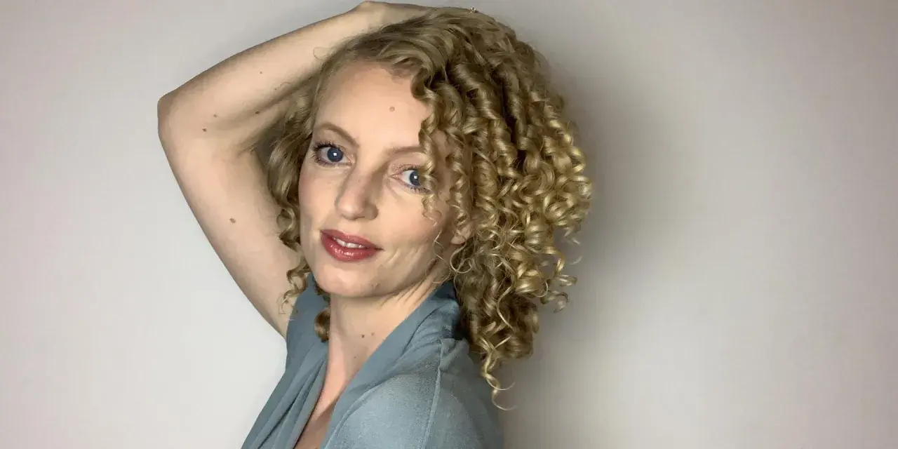 Frau mit blonden, voluminösen 3a locken, die ihren Arm über den Kopf hebt.