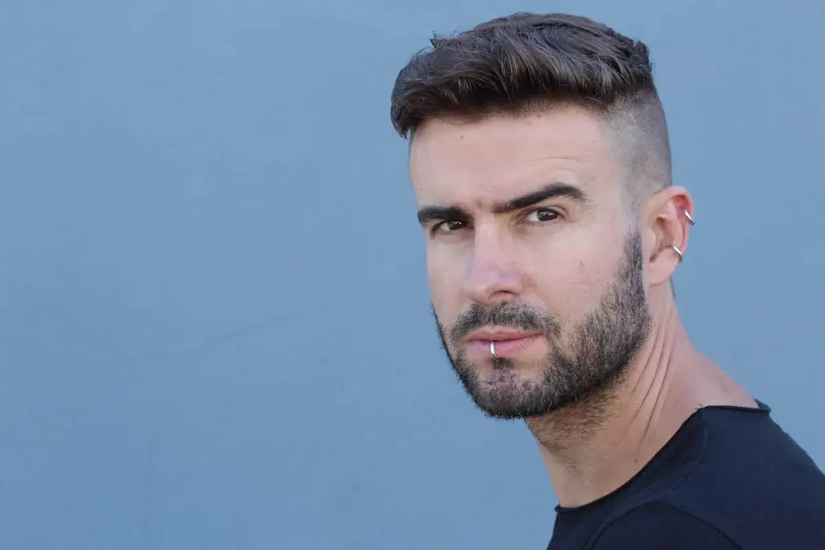 Junger Mann mit kurz geschorenen Seiten und Haare nach hinten, Bart und Lippenpiercing.