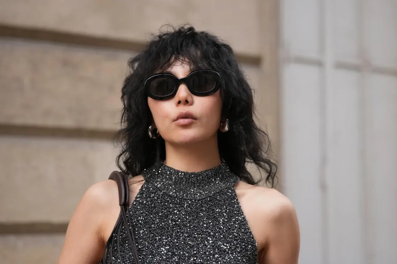 Frau mit schwarzem, gewelltem Haar im **Wolf Cut kurz**. Sie trägt eine glitzernde Top und eine Sonnenbrille.