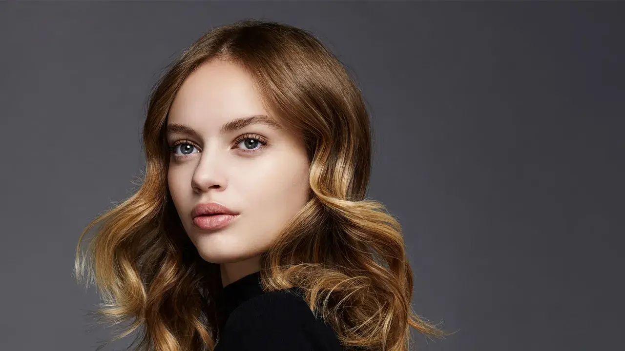 Porträt einer jungen Frau mit welligem, dunkelblondem Balayage-Haar, das sanft um ihr Gesicht fällt.