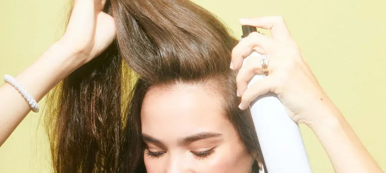 Frau sprüht Haarspray auf ihr Haar. Macht Haarspray die Haare kaputt? Die Antwort liegt im richtigen Gebrauch.