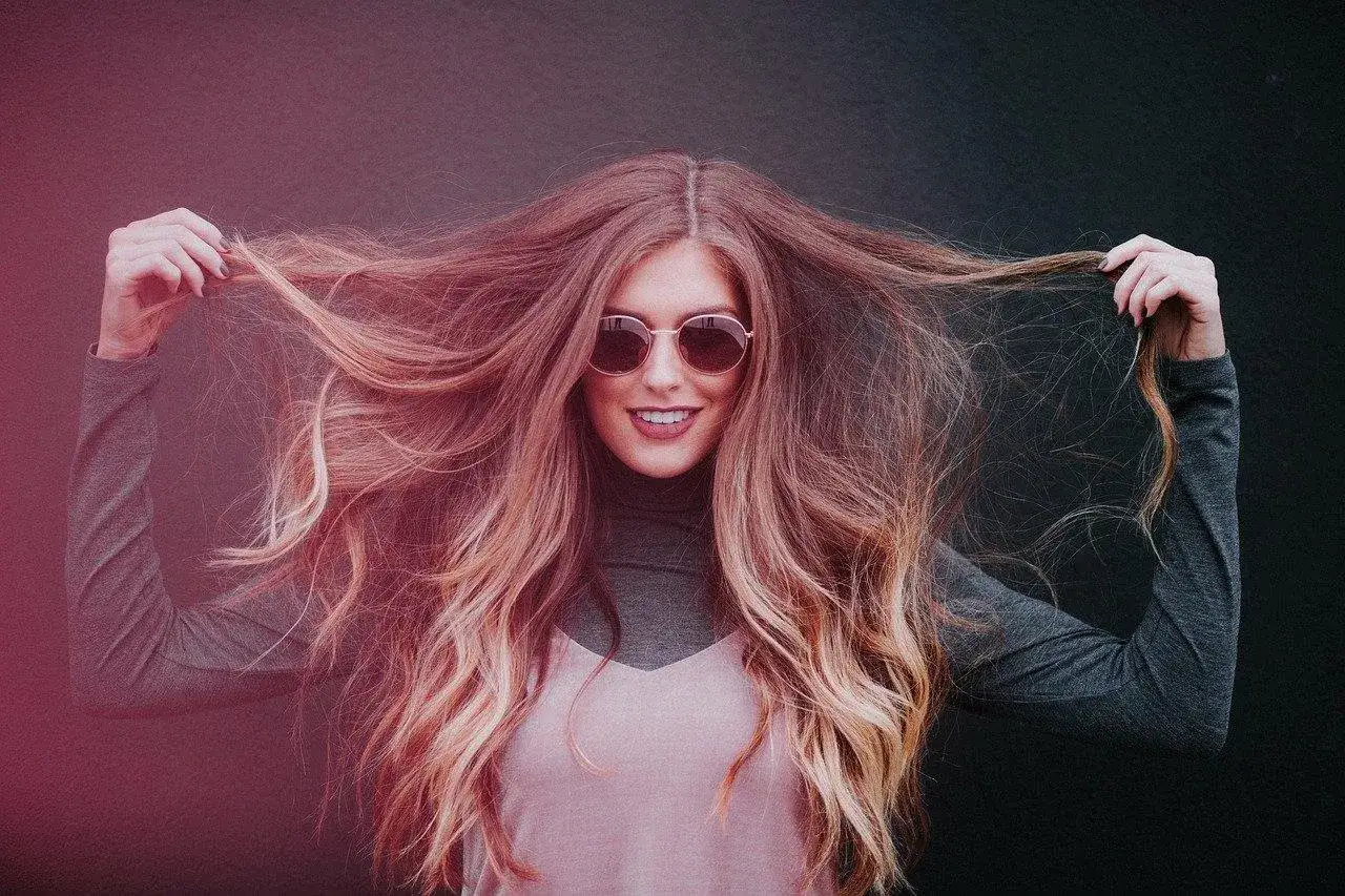 Frau mit langen, dicken Haaren, die sie in die Luft wirft. Sie trägt eine Sonnenbrille und lächelt.