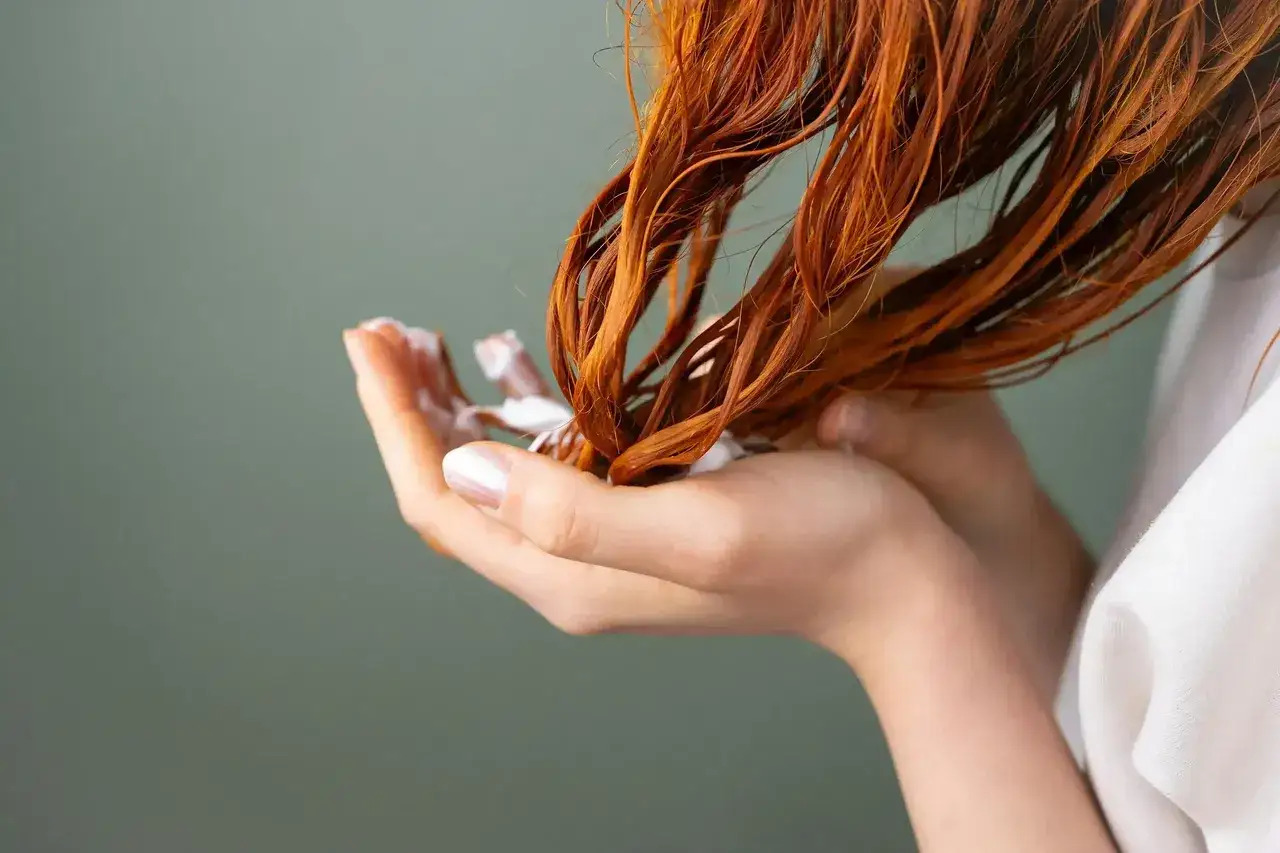 Hände mit weißer Creme, die in feuchtes, rotes Haar einmassiert wird. Eine wohltuende Haarkur für trockenes Haar.