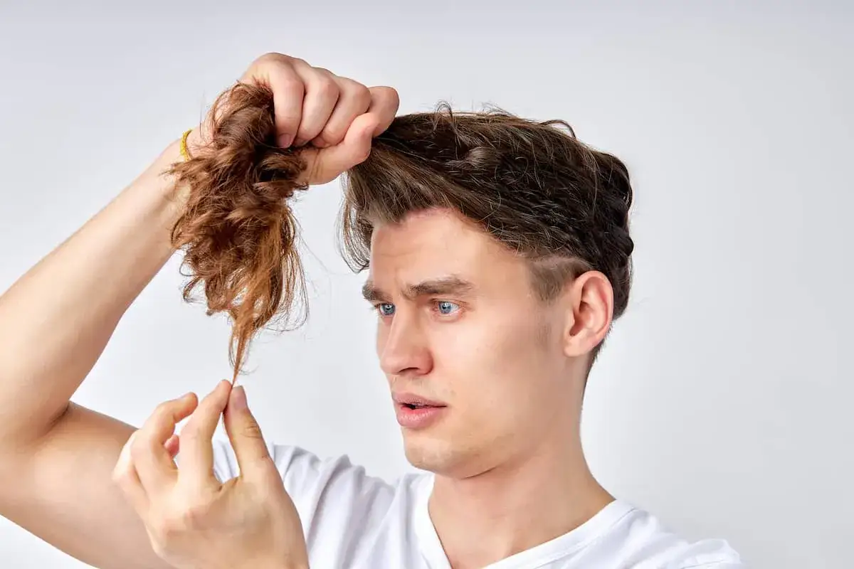 Mann mit blauen Augen betrachtet besorgt seine kaputten Haare, die er in der Hand hält.