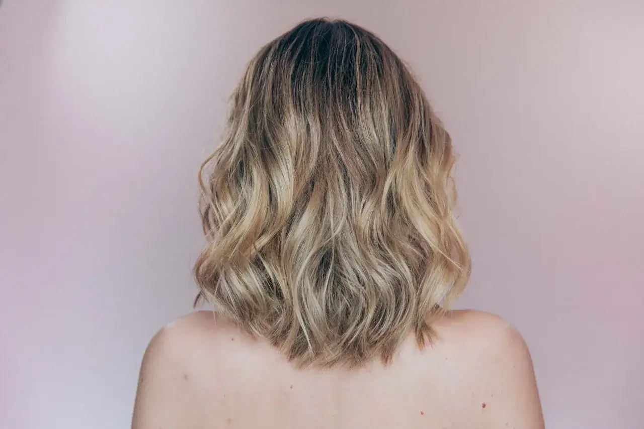 Rückenansicht einer Frau mit welligem, blondem Haar, das einen modernen, kalligrafischen Schnitt hat.