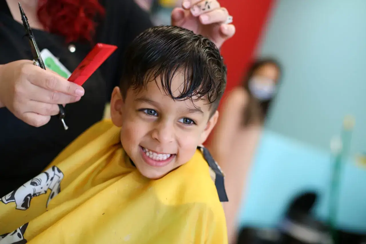 Ein Junge mit einem breiten Lächeln bekommt einen Haarschnitt. Die Friseurin hält eine Schere und einen Kamm bereit, um die Jungs Frisuren Namen zu kreieren.
