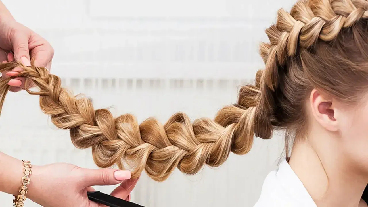 Eine Hand flechtet kunstvoll geflochtene Haare zu einem dicken Zopf. Die blonde M&auml;hne gl&auml;nzt im Licht.