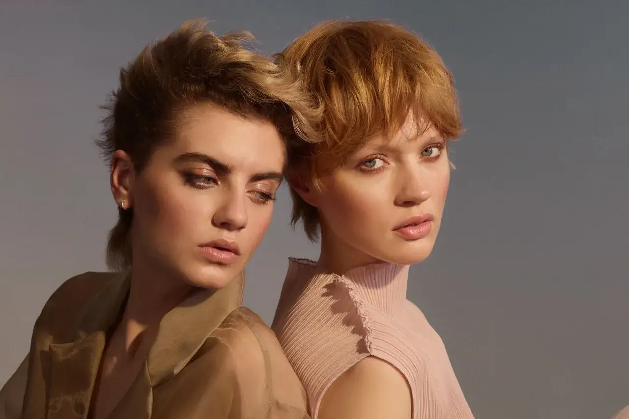 Zwei Frauen mit modernen Kurzhaarfrisuren, eine mit blondem, zerzaustem Haar, die andere mit rotem Pixie-Schnitt, posieren im sanften Licht.