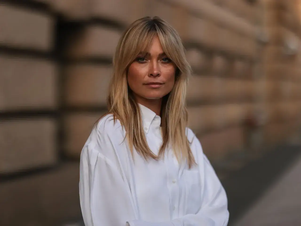 Frau mit blonden Haaren und wispy bangs, trägt ein weißes Hemd.