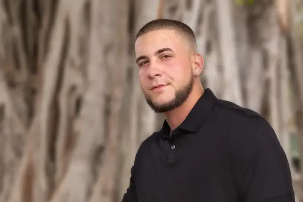 Junger Mann mit Bart und Buzz Cut 10mm, trägt schwarzes Poloshirt, steht vor unscharfem Baumstamm.
